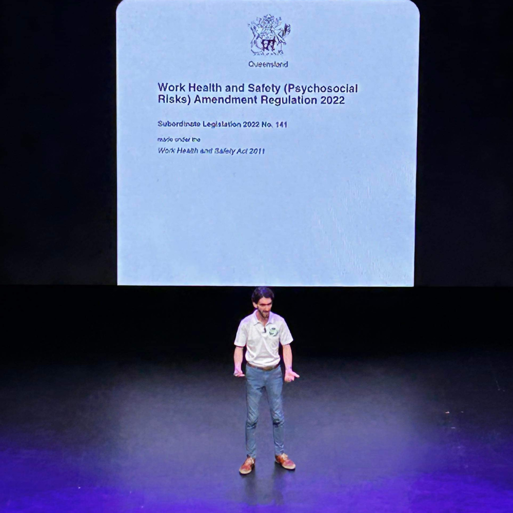 Presenter on stage beneath a slide about psychosocial safety regulations.